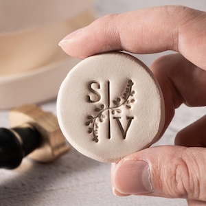 May include: A round, off-white wax seal stamp with the initials "S" and "V" separated by a floral design. The stamp is held by a person's fingers, with a gold-colored handle visible in the background. The stamp is for crafts and stationery.