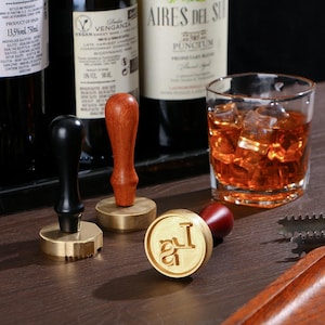 Op de afbeelding: Een verzameling messing waszegelstempels met houten handvatten, samen met een glas amberkleurige vloeistof met ijsblokjes. Wijnflessen staan op de achtergrond. De stempels hebben verschillende ontwerpen, waaronder een cirkelvormige met initialen.
