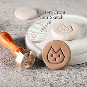 May include: A wooden-handled stamp with a brass head, alongside a brown clay stamp with a cat face design. The image also shows a sketch of the cat face and the text "Custom From Your Sketch".