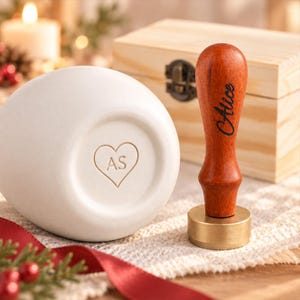 May include: A wax seal stamp with a reddish-brown wooden handle engraved with the name Alice and a brass circular base. Beside it sits a white ceramic object featuring an embossed heart shape containing the letters AS, with a wooden box behind.