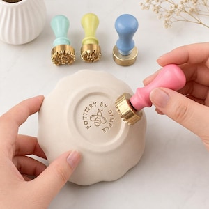 May include: A pink pottery stamp with a brass head is pressing into a scalloped ceramic dish, leaving a circular impression that reads POTTERY BY DIMPLE with a bee icon. Three additional stamps in mint, yellow, and blue are arranged nearby.