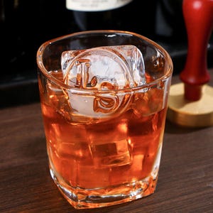 May include: A square glass filled with an amber-colored liquid and ice cubes. A large ice cube with the letters "LS" is in the drink. The glass sits on a dark wooden surface. A red object is in the background.