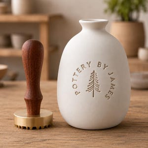 May include: A matte white ceramic vase with a narrow neck and a rounded body, featuring the embossed text POTTERY BY JAMES and a pine tree icon. Beside it stands a brass stamp with a dark brown wooden handle used for clay impressions.