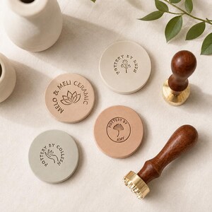 May include: Four circular clay discs with custom embossed logos, including designs featuring a lotus, a flower, a ginkgo leaf, and a hand. Two wooden-handled brass stamps are present, one resting on its side and one standing upright.