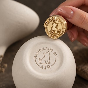 Może przedstawiać: Okrągła mosiężna pieczątka z sylwetką kota i napisem HANDMADE BY AJR, umieszczona nad pasującym okrągłym odciskiem na matowej, kremowobiałej powierzchni ceramicznej. Pieczątka posiada ciemny drewniany uchwyt i złote wykończenie.