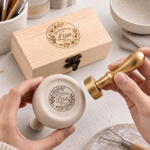 May include: A brass pottery stamp with a smooth handle and a circular head featuring a floral design and the text "POTTERY MADE BY Lisa". The stamp is shown next to a rectangular wooden storage box with a matching floral logo engraved on the lid.