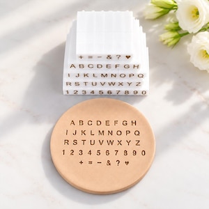 May include: A set of white square stamps featuring letters, numbers, and symbols, including a heart, arranged in rows. Below the stamps is a circular tan clay disc embossed with the same alphabet, numbers, and symbols in a matching layout.