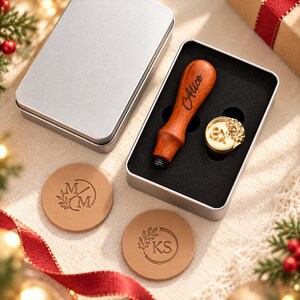 May include: A wax seal stamp set featuring a wooden handle engraved with the name Alice, a brass stamp head with an ampersand and leaf design, and two round tan leather coasters embossed with the initials MM and KS, all housed in a silver tin.