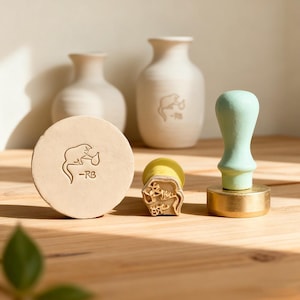 May include: A selection of craft items, including a round stamp, a rectangular stamp, and a stamp with a pale green handle. The stamps feature a rat design and the initials "-RB". Two off-white vases are in the background.