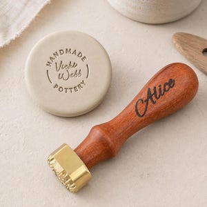 Könnte beinhalten: Ein Töpferstempel mit Holzgriff, auf dem der Name Alice in schwarzer Schreibschrift eingraviert ist, befestigt an einem Messingkopf. Daneben liegt eine kreisförmige, hellbeige Tonscheibe mit der Prägung Handmade Vicki Webb Pottery.