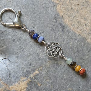 May include: A silver-toned metal keychain featuring a lobster clasp, a central silver-toned flower of life charm, and seven faceted gemstone beads in shades of dark purple, blue, light blue, pale green, brown, yellow, and reddish-orange.