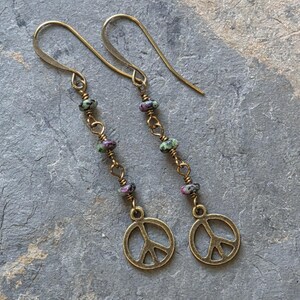 May include: A pair of dangle earrings featuring brass-toned metal hooks and chains. Each earring is accented with three small, multi-colored speckled beads in shades of green, black, and magenta, terminating in a circular peace sign charm.