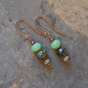 May include: A pair of handcrafted earrings featuring copper ear wires and a combination of green and speckled beads. The earrings have a drop design with faceted and round beads in shades of green and brown.