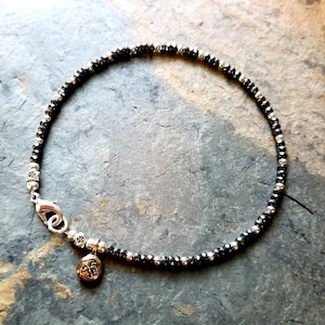 May include: Ankle bracelet featuring small, black and silver beads strung together. The bracelet has a silver clasp and a small charm with a decorative design. The jewelry is displayed on a textured, gray surface.