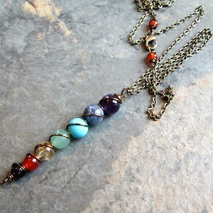 May include: A handmade necklace featuring a series of colorful gemstone beads, including amethyst, turquoise, and citrine, strung on a bronze-colored chain. The beads are wrapped in bronze wire, and the necklace has a lobster clasp closure. The design is inspired by chakra energy.