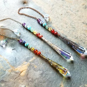 May include: Three crystal pendulum wands with colorful chakra beads. Each wand has a different metal chain and decorative tip. The beads are arranged in a rainbow pattern, transitioning from purple to red.