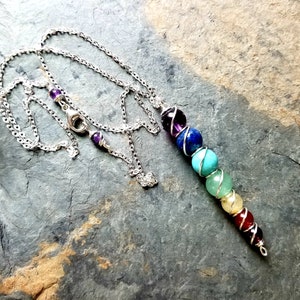 May include: A silver chain necklace with a pendant featuring seven colorful gemstone beads, representing the chakras. The beads are wrapped in silver wire. The necklace is displayed on a gray stone surface.