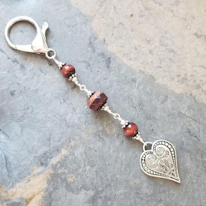 May include: A silver-toned keychain charm with a lobster clasp. The charm features a chain of red-brown beads and a decorative heart pendant. The heart has an ornate, swirling design.