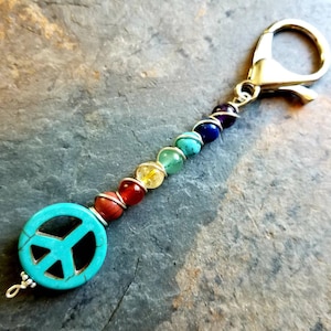 May include: A keychain featuring a turquoise peace sign charm and a strand of colorful beads representing the chakras. The beads are wrapped in silver wire and attached to a gold-colored clasp.