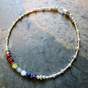 May include: Silver bracelet featuring a series of colorful gemstone beads representing the chakras. The bracelet has a delicate design with small silver beads and a secure clasp. The gemstones include red, orange, yellow, green, blue, indigo, and purple.
