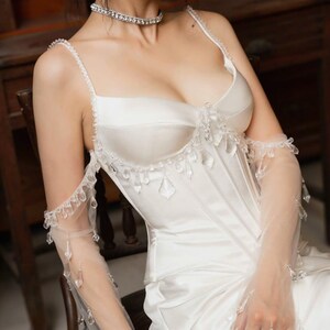 May include: A white satin corset-style top with delicate crystal embellishments and sheer, long gloves. The top features thin straps and a sweetheart neckline. The gloves are adorned with hanging crystals. A sparkling choker necklace completes the look.