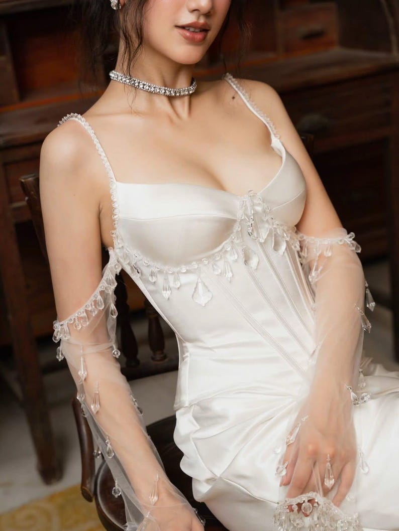 May include: A white satin corset-style top with delicate crystal embellishments and sheer, long gloves. The top features thin straps and a sweetheart neckline. The gloves are adorned with hanging crystals. A sparkling choker necklace completes the look.