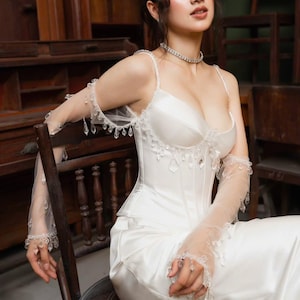 May include: A white satin wedding dress with a fitted bodice and a flowing skirt. The dress features delicate spaghetti straps and is adorned with crystal embellishments. Sheer, long gloves with crystal accents complete the elegant bridal look. The model is seated on a wooden chair.