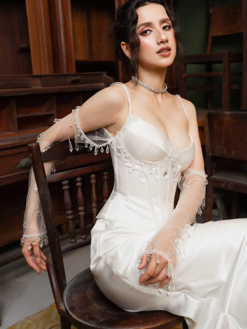 May include: A white satin wedding dress with a fitted bodice and a flowing skirt. The dress features delicate spaghetti straps and is adorned with crystal embellishments. Sheer, long gloves with crystal accents complete the elegant bridal look. The model is seated on a wooden chair.