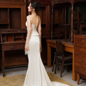May include: A white satin wedding dress with a corset bodice and a long train. The dress has spaghetti straps and delicate lace detailing on the sleeves. The bride is standing in a room with antique wooden furniture.