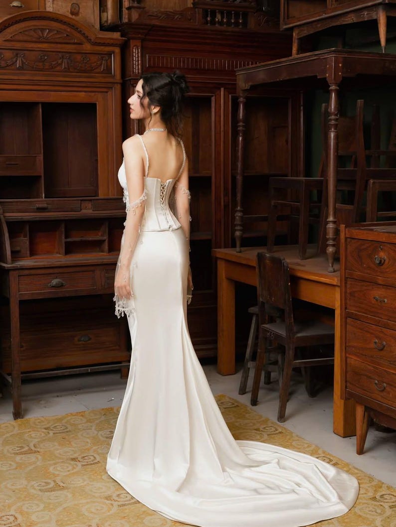May include: A white satin wedding dress with a corset bodice and a long train. The dress has spaghetti straps and delicate lace detailing on the sleeves. The bride is standing in a room with antique wooden furniture.