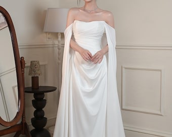 May include: A white off-the-shoulder gown with draped fabric and flowing sleeves. The dress has a fitted bodice and a floor-length skirt. The model is wearing a necklace and earrings. The background includes a mirror, a side table, and framed artwork.