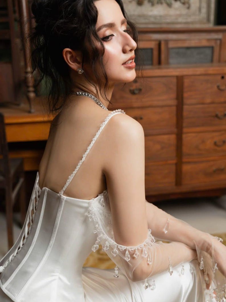 May include: A white satin dress with a gray corset-style bodice and delicate lace trim. The dress features thin straps and is paired with sheer gloves adorned with crystal accents. The woman is wearing a diamond necklace and earrings.