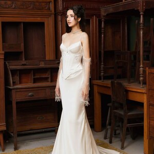 May include: A white satin wedding gown with a fitted bodice and a flowing skirt. The dress features delicate spaghetti straps and sheer, embellished sleeves. The bride is standing in a room with antique wooden furniture.
