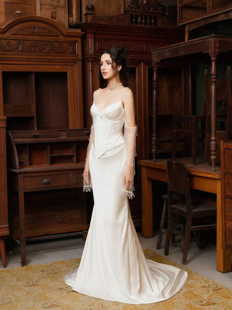 May include: A white satin wedding gown with a fitted bodice and a flowing skirt. The dress features delicate spaghetti straps and sheer, embellished sleeves. The bride is standing in a room with antique wooden furniture.