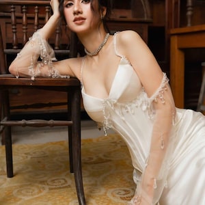 May include: A white satin gown with a sweetheart neckline and delicate beaded embellishments. The dress features spaghetti straps and a fitted bodice. Sheer, long gloves with dangling crystals add a touch of elegance. A diamond necklace completes the look.