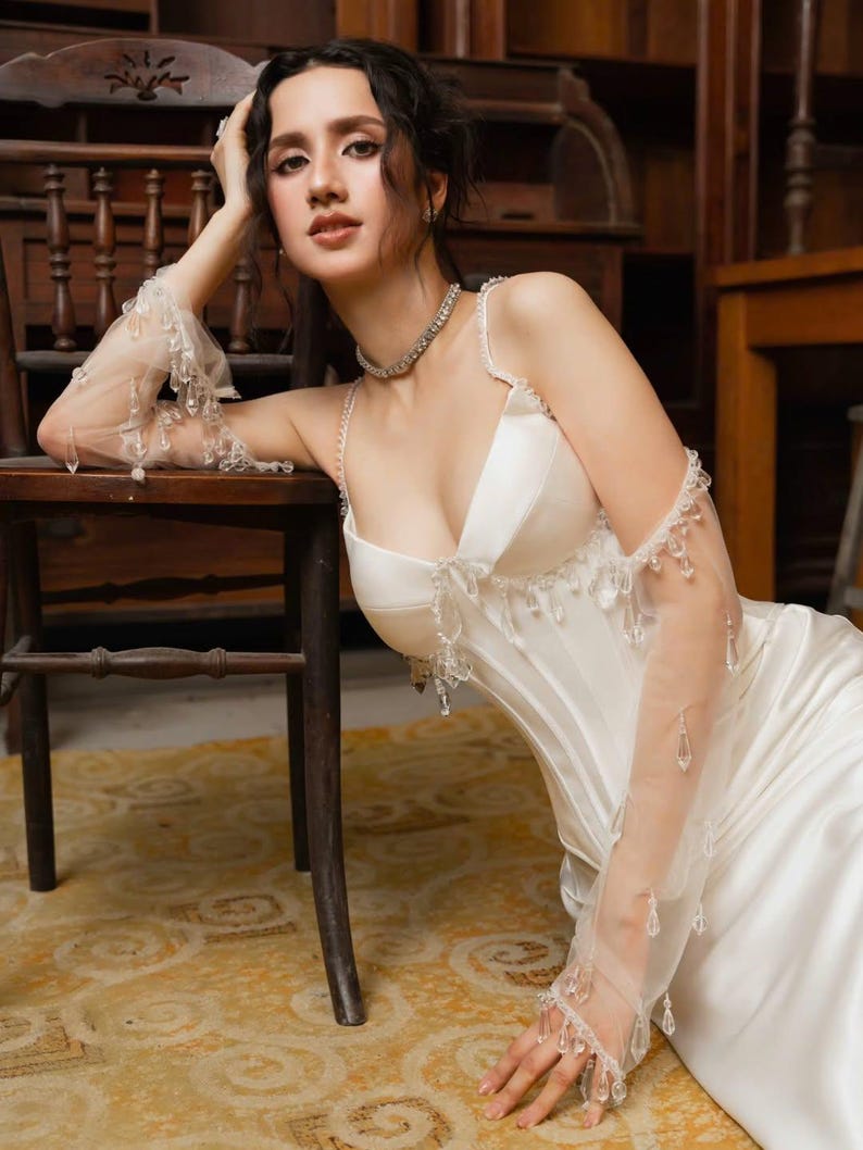 May include: A white satin gown with a sweetheart neckline and delicate beaded embellishments. The dress features spaghetti straps and a fitted bodice. Sheer, long gloves with dangling crystals add a touch of elegance. A diamond necklace completes the look.