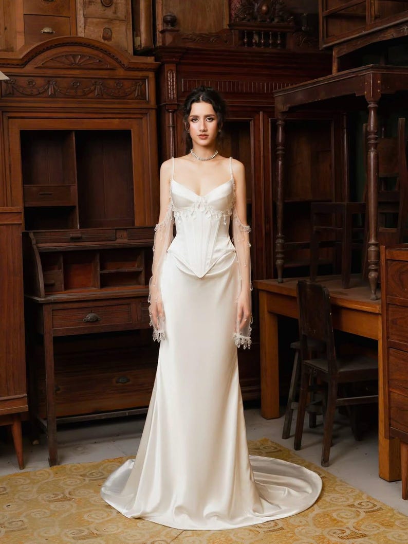 May include: A white satin wedding dress with a fitted bodice and a flowing skirt. The dress features delicate spaghetti straps and sheer sleeves with beaded accents. The bride is standing in a room with antique wooden furniture.