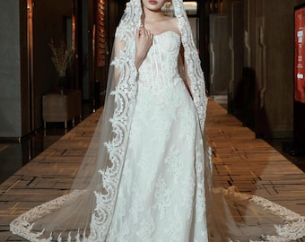 May include: A strapless white wedding dress with intricate lace detailing and a long, flowing train. The dress features a fitted bodice and a full skirt. A matching lace-trimmed veil drapes over the head and shoulders, creating an elegant bridal look.