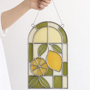 May include: Stained glass window panel featuring a lemon design. The panel has a rectangular shape with an arched top. The design includes a whole lemon, a lemon slice, and green leaves. The colors are yellow, green, and white.