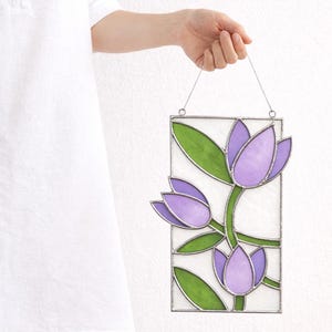 May include: A stained glass panel featuring a floral design with purple tulip-like flowers and green leaves. The panel is rectangular and held by a wire, showcasing a delicate and artistic piece of home decor.