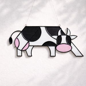 May include: A stained glass cow decoration with a black and white body, pink accents, and a black tail. The cow has a geometric design and is suspended by a thin wire. The background is a plain white surface.