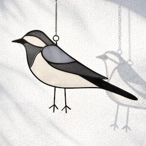 Puede incluir: Atrapasoles de pájaro de vidriera en tonos grises, blancos y negros, colgado de una cadena. El diseño del pájaro presenta piezas de vidrio segmentadas, creando una silueta aviar detallada. El atrapasoles proyecta una sombra en la pared.