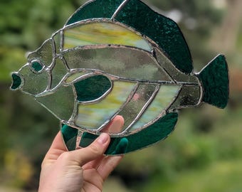 May include: Stained glass fish sculpture with a mix of green, yellow, and clear glass. The fish has a detailed design with fins and a tail. The glass is framed with a silver metal border. The sculpture is held up against a blurred green background.