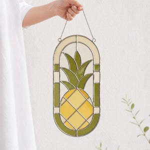 Puede incluir: Colgante de ventana de piña de vidrieras. El diseño presenta una piña amarilla con hojas verdes, enmarcada por un borde verde y crema. Una pieza decorativa para la decoración del hogar.