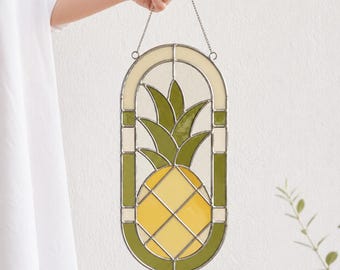 May include: Stained glass pineapple window hanging with a chain for display. The design features a yellow pineapple with green leaves, framed by a green and cream-colored border. A decorative piece for home decor.