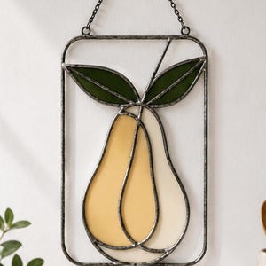 May include: A rectangular stained glass wall hanging featuring a pear design with two overlapping yellow and cream-colored glass segments and two dark green glass leaves, all encased in a dark metal frame with a matching hanging chain.