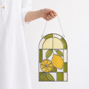 Puede incluir: Panel de ventana de vidrieras con diseño de limón. El panel tiene un borde a cuadros verde y blanco, con un limón y hojas amarillas y verdes en el centro. Se sujeta con una cadena plateada.