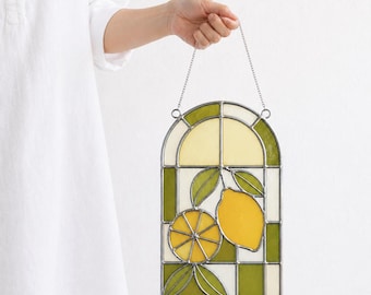 May include: Stained glass window panel featuring a lemon design. The panel has a green and white checkered border, with a yellow and green lemon and leaves in the center. The panel is held by a silver chain.