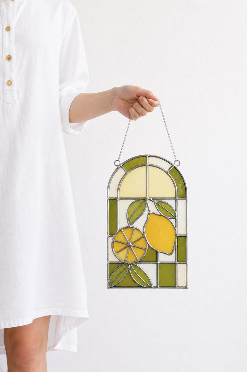 May include: Stained glass window panel featuring a lemon design. The panel has a green and white checkered border, with a yellow and green lemon and leaves in the center. The panel is held by a silver chain.