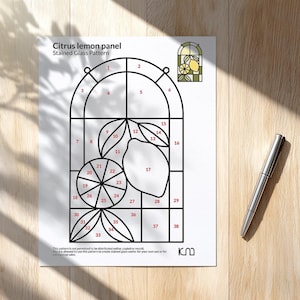 May include: A printed pattern for a citrus lemon stained glass panel. The design features a lemon and leaves within a window frame, with numbered sections for glass cutting. A silver pen rests on the wooden surface.
